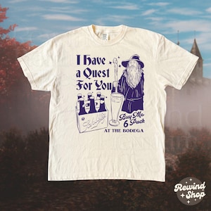 May include: Cream-colored t-shirt with a purple graphic. The graphic features a wizard holding a staff and a pint of beer, with text that reads "I Have a Quest For You" and "Buy Me 6 Pack At The Bodega".