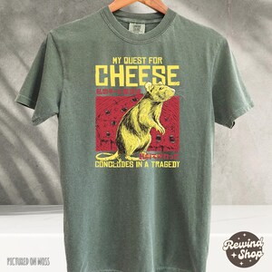 Op de afbeelding: Een groen T-shirt met een afbeelding van een rat en de tekst "My Quest for Cheese Concludes in a Tragedy".