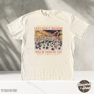 May include: Ivory-colored t-shirt featuring a graphic with a red and black spider-like creature over a field of eyeballs. The text reads "I HAVE MADE IT AWKWARD" and "I WILL BE TAKING MY LIFE" in English and Japanese.