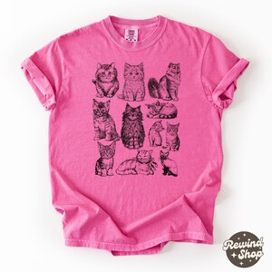 K&ouml;nnte beinhalten: Ein pinkfarbenes T-Shirt mit Illustrationen von schwarzen und wei&szlig;en Katzen. Die Katzen sind in verschiedenen Posen dargestellt, sitzend, liegend und stehend. Das T-Shirt hat hochgekrempelte &Auml;rmel und einen Rundhalsausschnitt. Der Markenname "Comfort Colors" ist auf dem Etikett zu sehen.