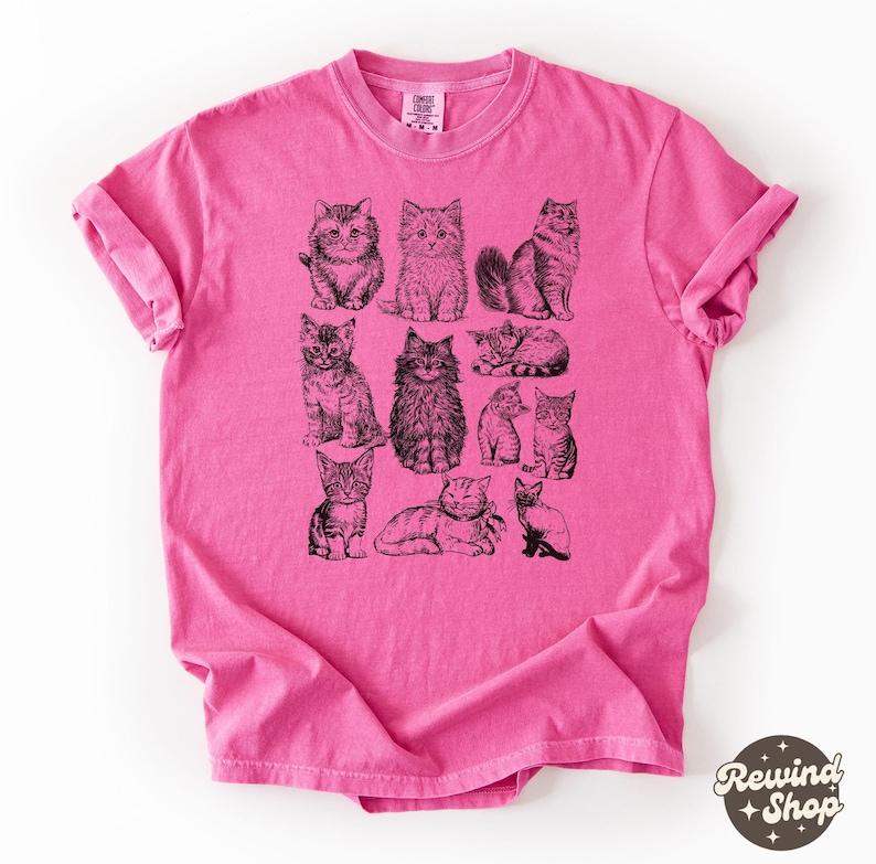 K&ouml;nnte beinhalten: Ein pinkfarbenes T-Shirt mit Illustrationen von schwarzen und wei&szlig;en Katzen. Die Katzen sind in verschiedenen Posen dargestellt, sitzend, liegend und stehend. Das T-Shirt hat hochgekrempelte &Auml;rmel und einen Rundhalsausschnitt. Der Markenname "Comfort Colors" ist auf dem Etikett zu sehen.