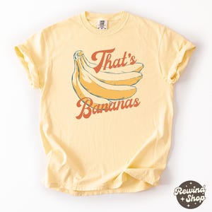 May include: A light yellow t-shirt featuring a bunch of bananas with the text "That's Bananas" in orange and brown. The shirt has a relaxed fit and rolled sleeves, perfect for casual wear.