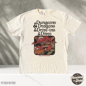 May include: Ivory-colored t-shirt with the text "Dungeons & Dragons & Drive-ins & Dives" in a retro font. The shirt features a graphic of a red monster holding a plate of food. The phrase "Escape From Flavortown" is below the graphic.