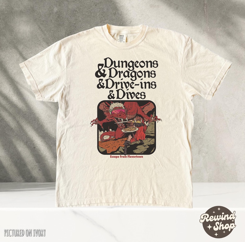 Może przedstawiać: Kremowa koszulka z napisem "Dungeons & Dragons & Drive-ins & Dives" w stylu retro. Koszulka posiada grafikę czerwonego potwora trzymającego talerz z jedzeniem. Poniżej grafiki znajduje się napis "Escape From Flavortown".