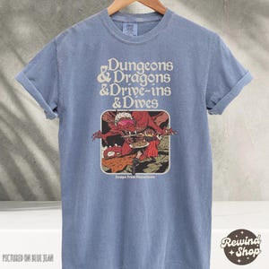Może przedstawiać: Niebieska koszulka z podwiniętymi rękawami z napisem "Dungeons & Dragons & Drive-ins & Dives". Grafika przedstawia czerwonego smoka i osobę. Poniżej grafiki znajduje się napis "Escape From Flavortown". Koszulka wisi na drewnianym wieszaku.