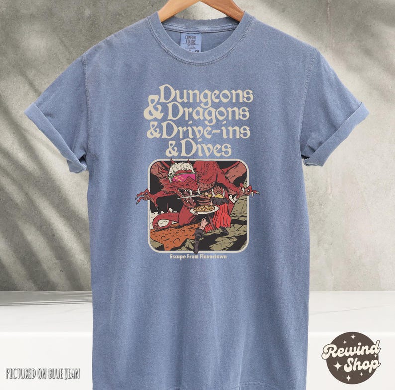 Może przedstawiać: Niebieska koszulka z podwiniętymi rękawami z napisem "Dungeons & Dragons & Drive-ins & Dives". Grafika przedstawia czerwonego smoka i osobę. Poniżej grafiki znajduje się napis "Escape From Flavortown". Koszulka wisi na drewnianym wieszaku.