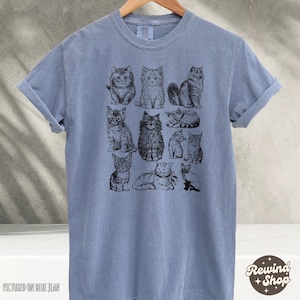 K&ouml;nnte beinhalten: Blaues T-Shirt mit Illustrationen von schwarzen und wei&szlig;en Katzen. Die Katzen sind in verschiedenen Posen dargestellt, sitzend, liegend und stehend. Das T-Shirt h&auml;ngt an einem Holzb&uuml;gel.