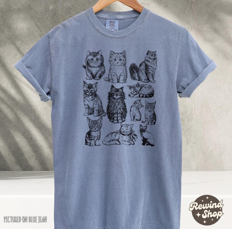 K&ouml;nnte beinhalten: Blaues T-Shirt mit Illustrationen von schwarzen und wei&szlig;en Katzen. Die Katzen sind in verschiedenen Posen dargestellt, sitzend, liegend und stehend. Das T-Shirt h&auml;ngt an einem Holzb&uuml;gel.
