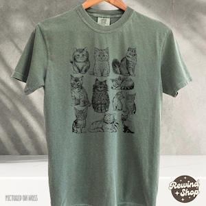 K&ouml;nnte beinhalten: Ein moosgr&uuml;nes T-Shirt mit einer Schwarz-Wei&szlig;-Illustration verschiedener Katzen. Die Katzen sind in verschiedenen Posen dargestellt. Das Shirt hat den Text "Comfort Colors" am Kragen und "Rewind Shop" unten rechts. Der Text "Pictured on Moss" steht unten links.
