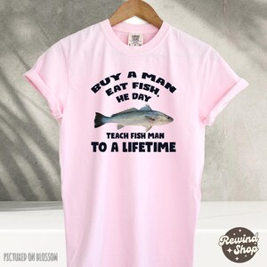May include: Pink t-shirt with a graphic design that says "Buy a man eat fish, he day teach fish man to a lifetime". The graphic also includes a drawing of a fish.