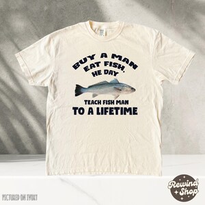 May include: A cream-colored t-shirt with a black graphic that reads "Buy a man eat fish, he day teach fish man to a lifetime". The graphic also includes a black and white illustration of a fish.