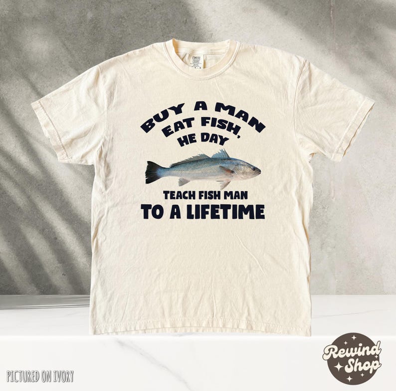 May include: A cream-colored t-shirt with a black graphic that reads "Buy a man eat fish, he day teach fish man to a lifetime". The graphic also includes a black and white illustration of a fish.