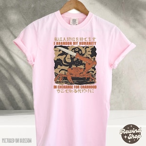 May include: A light pink t-shirt with a graphic design featuring a crab holding a knife, with Japanese and English text. The text reads "I abandon my humanity in exchange for crabhood".