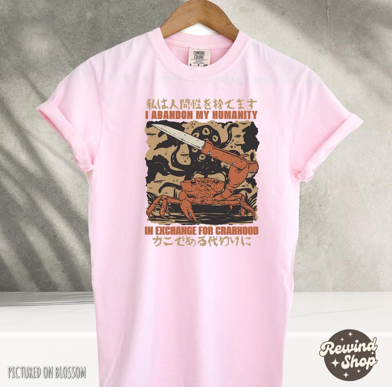 May include: A light pink t-shirt with a graphic design featuring a crab holding a knife, with Japanese and English text. The text reads "I abandon my humanity in exchange for crabhood".