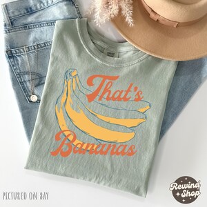 May include: A light green t-shirt with a banana graphic and the text "That's Bananas" in orange. The shirt is folded on top of blue jeans, with a gold necklace and a tan hat visible.