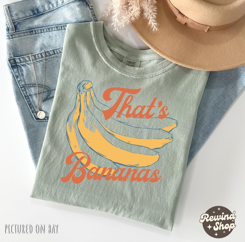 May include: A light green t-shirt with a banana graphic and the text "That's Bananas" in orange. The shirt is folded on top of blue jeans, with a gold necklace and a tan hat visible.
