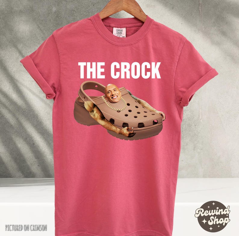 May include: A red t-shirt with the text "THE CROCK" printed in white. A brown and black Croc shoe is pictured with a person's face in the shoe. The person's face is Dwayne "The Rock" Johnson.