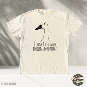 May include: A cream-colored t-shirt with a black and white illustration of a goose. The text "I think I will cause problems on purpose" is printed on the shirt.