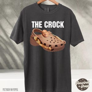 May include: A dark gray t-shirt with the text "THE CROCK" printed in white. The shirt features a brown and black Croc shoe with a picture of Dwayne "The Rock" Johnson's face on the shoe.