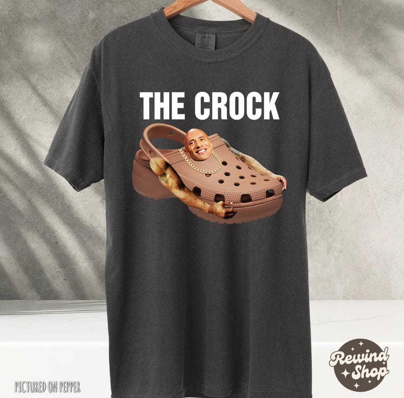 May include: A dark gray t-shirt with the text "THE CROCK" printed in white. The shirt features a brown and black Croc shoe with a picture of Dwayne "The Rock" Johnson's face on the shoe.