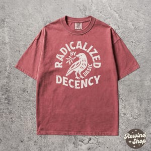 Puede incluir: Camiseta de color rosa polvoriento con cuello redondo y mangas cortas. En la parte delantera, un gráfico blanco de un pájaro con una ramita, rodeado por las palabras "RADICALIZED BY BASIC DECENCY". La camiseta tiene un corte holgado.