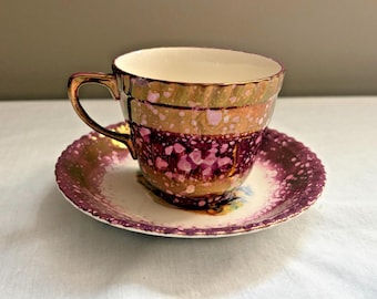 May include: A ceramic teacup and saucer set featuring a metallic gold and deep purple luster glaze with a speckled pattern. The teacup has a fluted rim and a curved handle, while the saucer has a scalloped edge and a matching purple speckled finish.