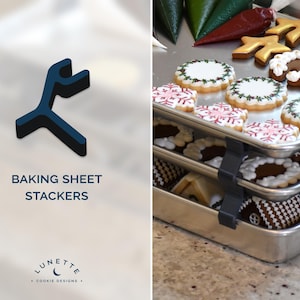 Puede incluir: Apiladores de bandejas para hornear, un juego de dos bandejas para hornear de metal con clips para sujetarlas. Las bandejas están apiladas con galletas encima, incluyendo hombres de jengibre, copos de nieve y otros diseños navideños.