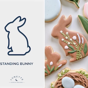 Pode incluir: Um cortador de biscoitos em forma de coelho em pé. O cortador é delineado em azul escuro sobre um fundo branco. O texto "STANDING BUNNY" está abaixo do cortador. O texto "LUNETTE COOKIE DESIGNS" está abaixo do texto "STANDING BUNNY".