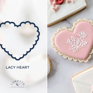 Puede incluir: Un cortador de galletas en forma de corazón calado azul marino y una galleta rosa decorada en forma de corazón con detalles florales blancos. La galleta tiene un borde festoneado. La imagen también muestra una galleta blanca decorada para parecer un sobre.