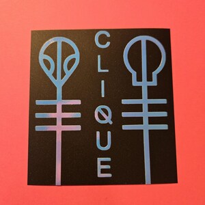 Pode incluir: Adesivo quadrado preto com dois s&iacute;mbolos estilizados e a palavra "CLIQUE" em azul claro. Os s&iacute;mbolos t&ecirc;m um gradiente azul e rosa. O fundo &eacute; vermelho liso.