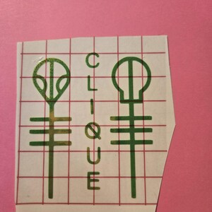 Pode incluir: Um autocolante com dois s&iacute;mbolos verdes e a palavra "CLIQUE" em verde. Os s&iacute;mbolos s&atilde;o estilizados, com linhas horizontais e elementos circulares. O autocolante est&aacute; sobre um suporte quadriculado, sobre um fundo rosa.