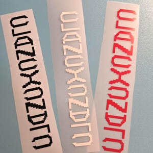 Peut inclure: Trois autocollants rectangulaires avec le texte "OZNOXDRO" dans une police pixélisée. Les autocollants sont blancs, noirs et rouges. Les autocollants sont sur une surface bleu clair.