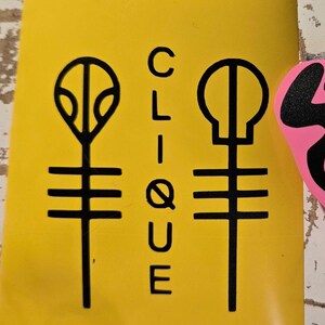 Pode incluir: Um autocolante amarelo com gr&aacute;ficos pretos e a palavra "CLIQUE" em texto vertical. O autocolante apresenta dois s&iacute;mbolos estilizados, um que se assemelha a uma planta e o outro a uma caveira, ambos com linhas horizontais.