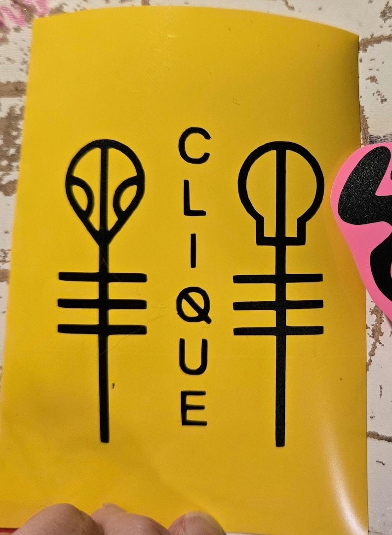 Pode incluir: Um autocolante amarelo com gr&aacute;ficos pretos e a palavra "CLIQUE" em texto vertical. O autocolante apresenta dois s&iacute;mbolos estilizados, um que se assemelha a uma planta e o outro a uma caveira, ambos com linhas horizontais.