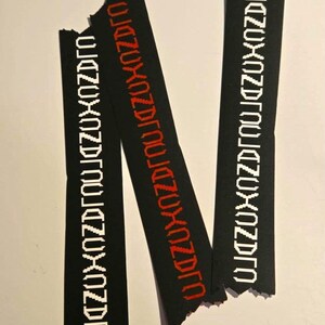 Könnte beinhalten: Drei schwarze Stoffstreifen mit zerfransten Kanten, die jeweils ein sich wiederholendes Muster aus weißem oder rotem Text aufweisen. Der Text scheint ein stilisiertes Design zu sein. Die Streifen sind vor einem weißen Hintergrund angeordnet.