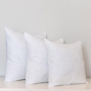 Puede incluir: Tres almohadas cuadradas blancas dispuestas sobre una superficie blanca contra un fondo gris claro. Las almohadas están rellenas y tienen un aspecto suave y esponjoso. Las almohadas son de diferentes tamaños.