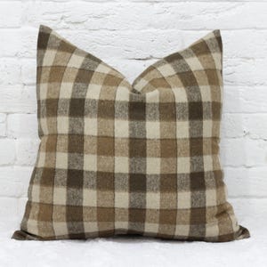 May include: A square decorative pillow with a brown and beige plaid pattern. The pillow has a soft, textured appearance and is placed against a white brick wall. The pillow is a home decor item.