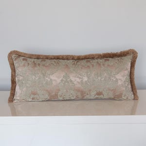 Puede incluir: Un cojín rectangular decorativo con un estampado damasco en tonos rosa y verde. El cojín tiene un borde con flecos marrones. Se encuentra sobre una superficie blanca.