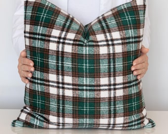 Puede incluir: Un gran cojín cuadrado con un estampado de cuadros verdes, marrones y blancos. El cojín está sostenido por una persona que lleva una camisa blanca. El cojín es un elemento decorativo para una cama o un sofá.