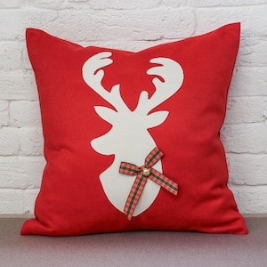 May include: A square red scatter cushion featuring a large, white, central silhouette of a reindeer head. The reindeer is decorated with a small, ornamental bow made of green and red tartan ribbon, which features a tiny metallic jingle bell at its centre.