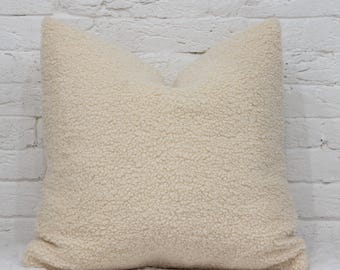 May include: A square, cream-colored pillow with a textured, plush surface. The pillow is made of a soft, fuzzy material and is set against a white brick wall.
