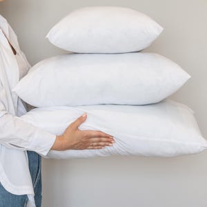 Op de afbeelding: Drie witte kussens op elkaar gestapeld. De kussens zijn rechthoekig en gevuld met een zacht materiaal. Een persoon in een wit overhemd en een blauwe spijkerbroek houdt de kussens vast. De kussens zijn een veelvoorkomend slaapkamerartikel.