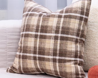 May include: A square decorative pillow with a brown, tan, and white plaid pattern. The pillow has a soft, textured appearance and is placed on a bed with other pillows. The pillow is a neutral color and would complement a variety of home decor styles.