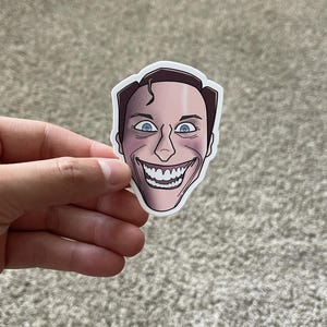 May include: A die-cut vinyl sticker featuring a stylized illustration of a human face with pale skin, dark hair, wide blue eyes, and an exaggerated, broad grin showing white teeth. The sticker has a white border and a smooth, matte finish.