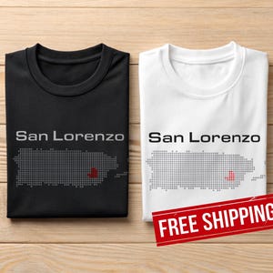 May include: Two folded t-shirts, one black and one white, with the text "San Lorenzo" above a dotted outline of Puerto Rico. The map has a red heart marking a location. The shirts are on a wooden surface.