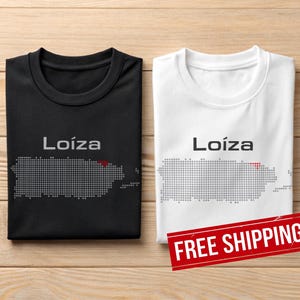 May include: Two t-shirts, one black and one white, with the word "Loíza" and a dotted map of Puerto Rico. The map has a red dot. The shirts are on a wooden surface.