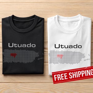 May include: Two t-shirts, one black and one white, each with the word "Utuado" printed above a dotted outline of Puerto Rico. The map has a red heart marking a location. The shirts are folded on a wooden surface.