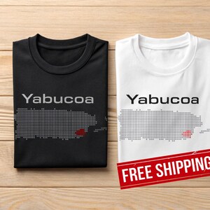 May include: Two t-shirts, one black and one white, with the word "Yabucoa" above a pixelated map of Puerto Rico. The map is grey with a red section. The black shirt is folded, and the white shirt is unfolded.