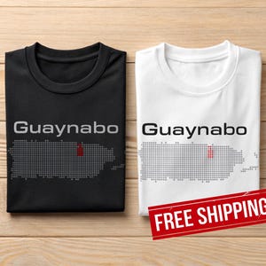 May include: Two folded t-shirts, one black and one white, with the word "Guaynabo" above a dotted outline of Puerto Rico. The black shirt has a grey outline, and the white shirt has a black outline. A red dot marks a location on the island.