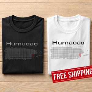 May include: Two t-shirts, one black and one white, with the word "Humacao" printed above a dotted outline of Puerto Rico. The black shirt has a red dot on the map. The white shirt has a red dot on the map.